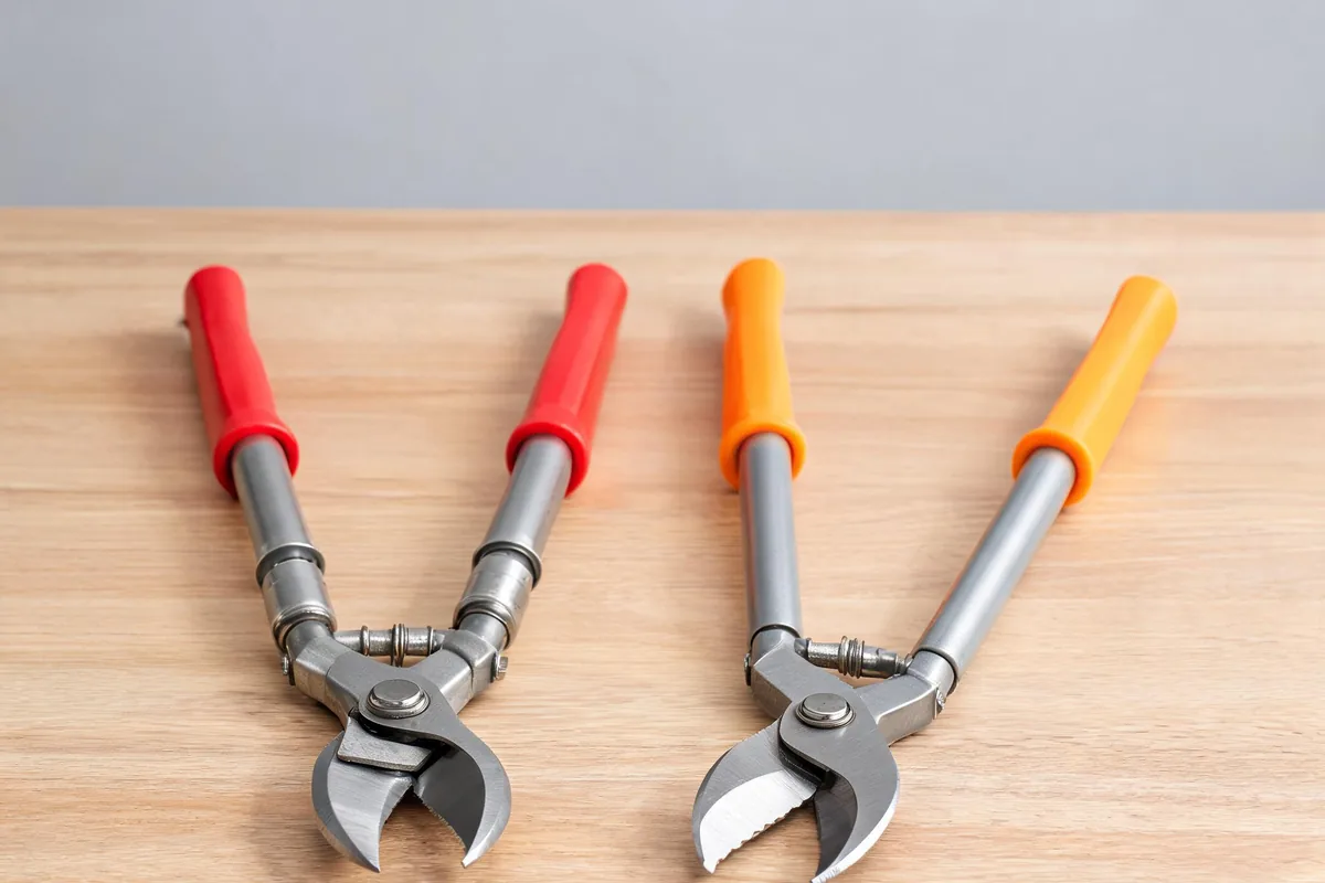 Two long-handled bypass loppers with red and orange grips on a wooden bench, one telescoping, showing curved cutting heads
