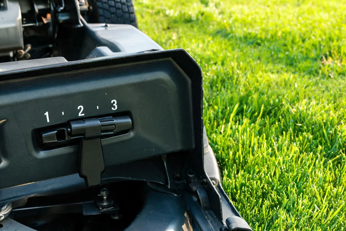 Walk-behind rotary lawn mower deck showing the height adjustment lever beside a freshly cut green lawn